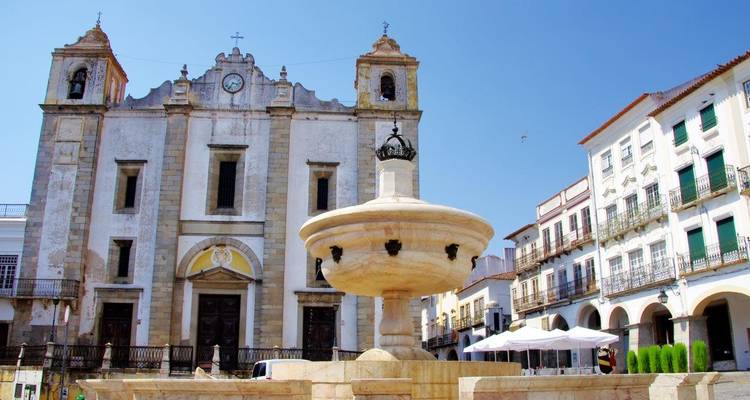 Steinerne Kirchenfassade und zentraler Brunnen auf einem sonnigen Platz in Évoras historischem Zentrum.