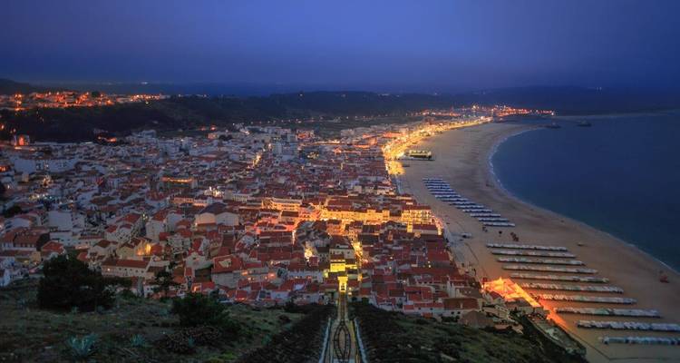Nachtansicht über die beleuchtete Küstenstadt und den weitläufigen Strand von Nazaré.