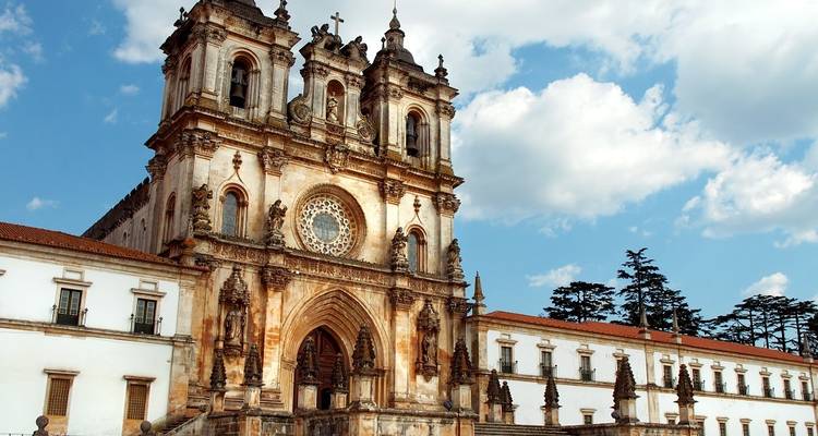 Gotische Fassade des Klosters von Alcobaça, beleuchtet von warmem Sonnenlicht mit flauschigen Wolken darüber.
