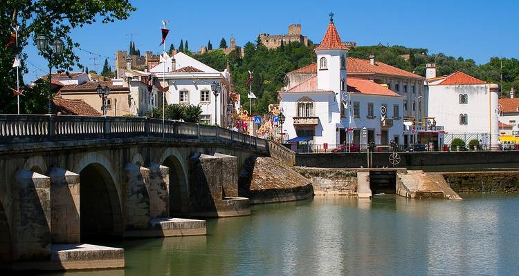 Steinbrücke über einen ruhigen Fluss mit weißgetünchten Gebäuden und einer Burganlage auf dem Hügel in Tomar.