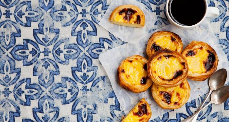 Draufsicht auf portugiesische Puddingtörtchen und Kaffee auf verzierten blauen Fliesen.