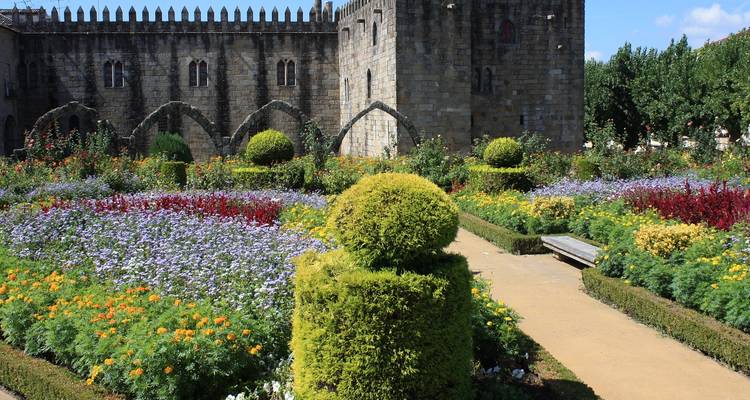Lebendige Blumenbeete und gepflegte Hecken im Garten von Santa Bárbara neben mittelalterlichen Steinmauern.