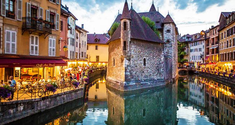 Bâtiment historique en pierre au milieu d'un canal illuminé au crépuscule avec des terrasses animées.
