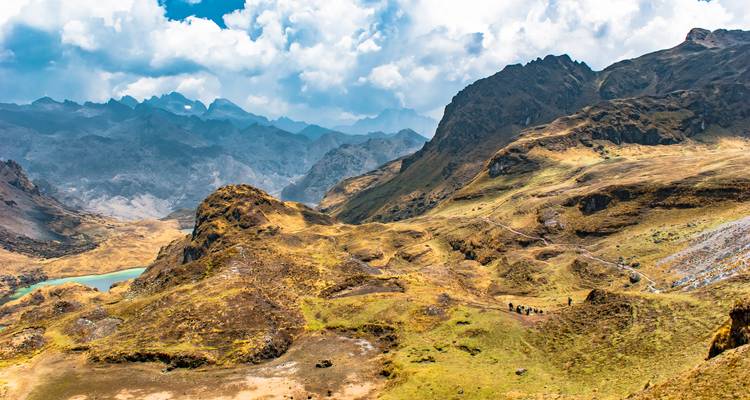Vue panoramique des Andes avec des collines dorées ondulantes, un lac turquoise et des pics dentelés sous des nuages éclatants.