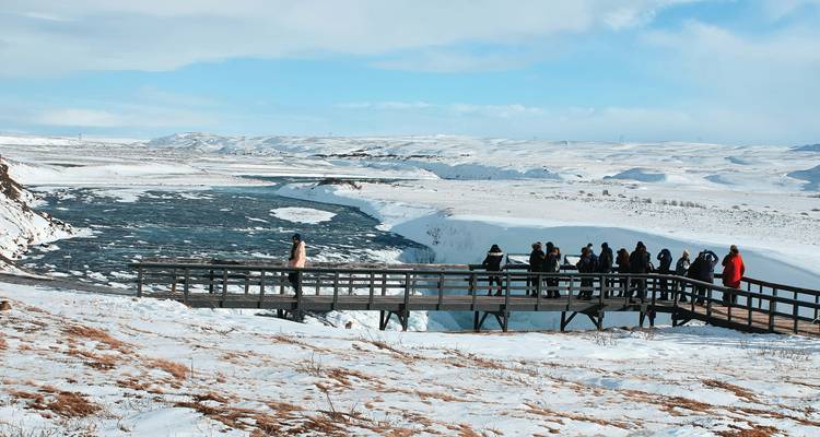 Tour group views frozen Gullfoss waterfall from a wooden platform in a snowy Icelandic landscape.