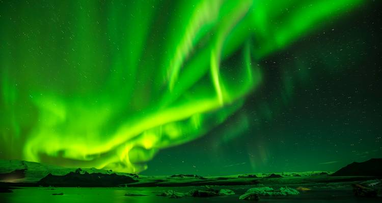 Brilliant green Northern Lights swirl across a starry Icelandic night sky above snowy peaks.