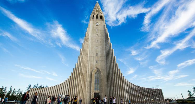 The soaring concrete spire of Hallgrímskirkja church reaches into a sky streaked with white clouds as visitors gather below.
