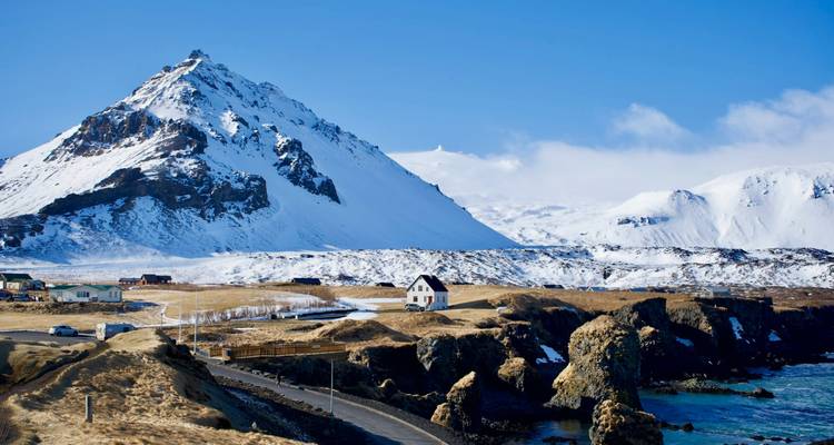 A snow-capped mountain rises above a rugged coastline dotted with a solitary white house and winding road.