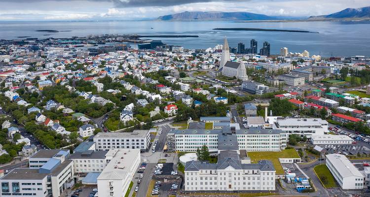 An aerial panorama of Reykjavik shows colorful buildings, Hallgrímskirkja and distant mountains beside the sea.