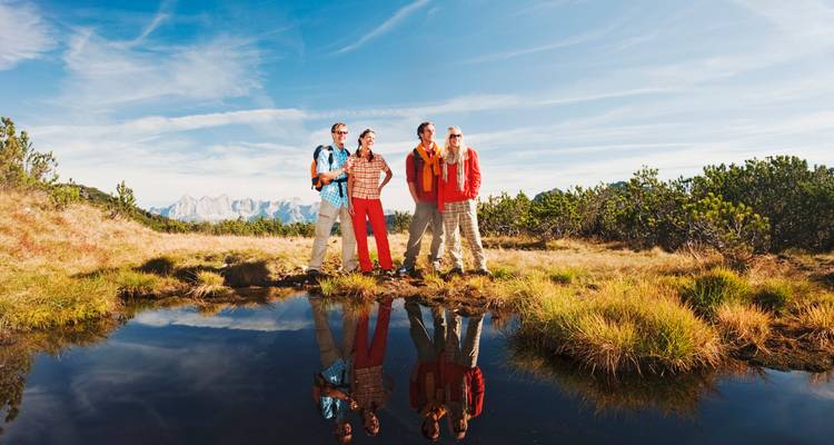 Cuatro excursionistas con equipo outdoor brillante están parados junto a un lago alpino reflectante en un día soleado, con picos de montaña escarpados en el horizonte detrás de ellos.