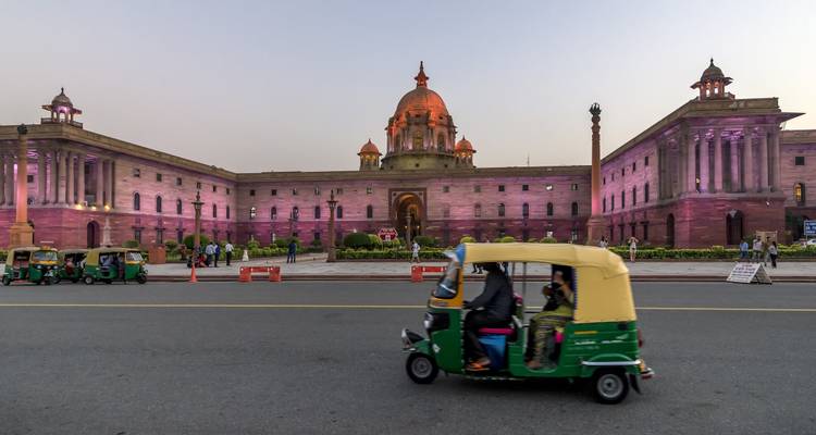 Personas caminando frente a un edificio gubernamental iluminado con tuk-tuks pasando.