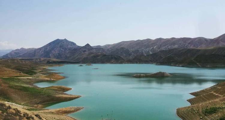 Un tranquilo embalse turquesa rodeado de colinas áridas y un pico montañoso distante bajo un cielo brumoso.