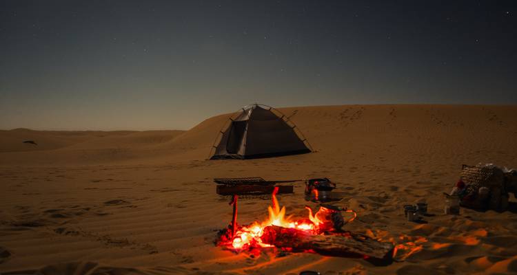 Feu de camp rougeoyant la nuit dans un désert avec une tente.