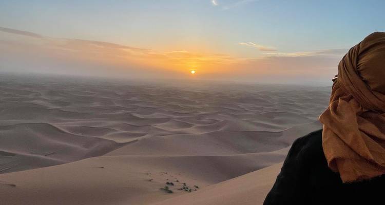 Voyageur enveloppé dans une écharpe observe le lever du soleil sur d'infinies dunes ondulées s'étendant jusqu'à l'horizon.