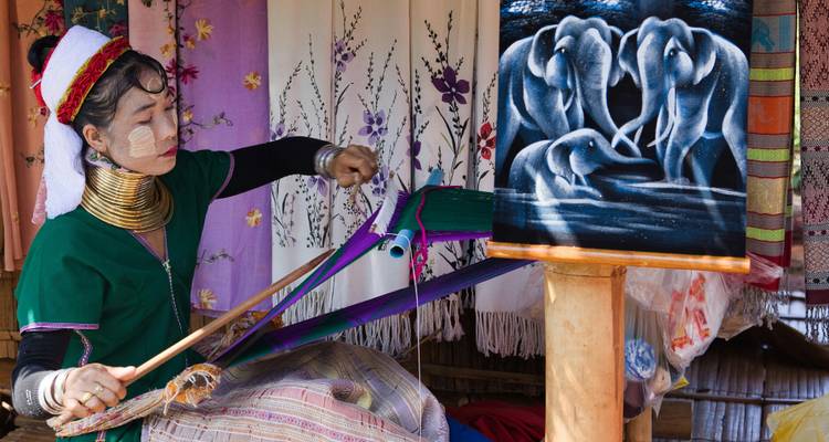 Una artesana Karen de cuello largo teje tela colorida junto a un cuadro de un elefante.