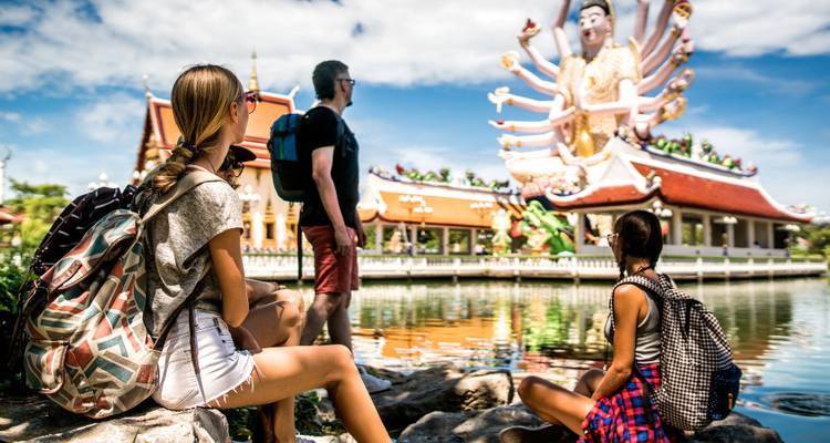 Tres turistas admiran una estatua ornamentada de múltiples brazos al otro lado de un estanque tranquilo.