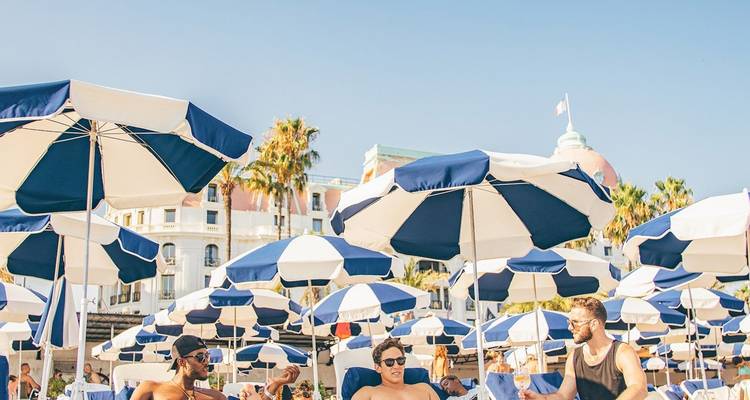Escena de club de playa soleado con filas de sombrillas azules, tumbonas y huéspedes relajados.