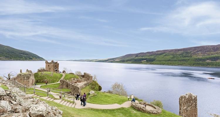 Besucher erkunden die grasbedeckten Ruinen von Urquhart Castle mit dem weitläufigen Loch Ness, der sich in die Ferne erstreckt