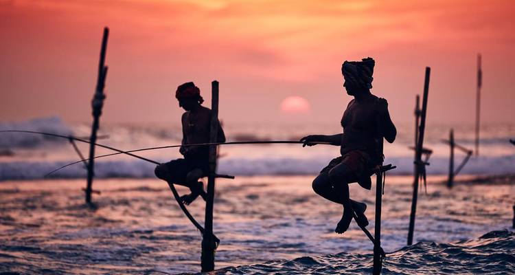 Silhouetted stilt fishermen cast lines against a vivid orange sunset over the ocean.