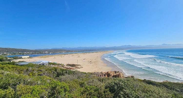 Large plage de sable avec des vagues douces et des montagnes lointaines sous un ciel bleu éclatant.