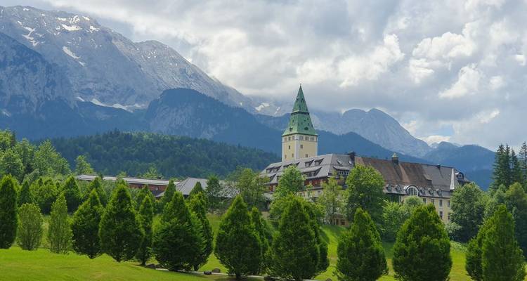 Un grand bâtiment avec une tour entouré de verdure luxuriante et de montagnes.