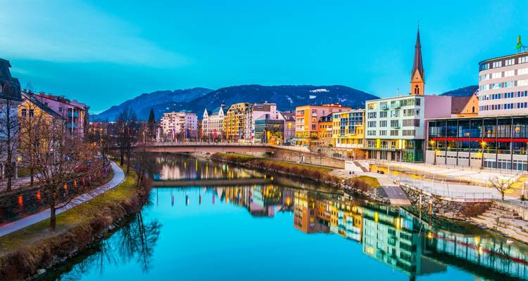 Vue colorée de la ville avec reflets sur la rivière et montagnes en arrière-plan au crépuscule.