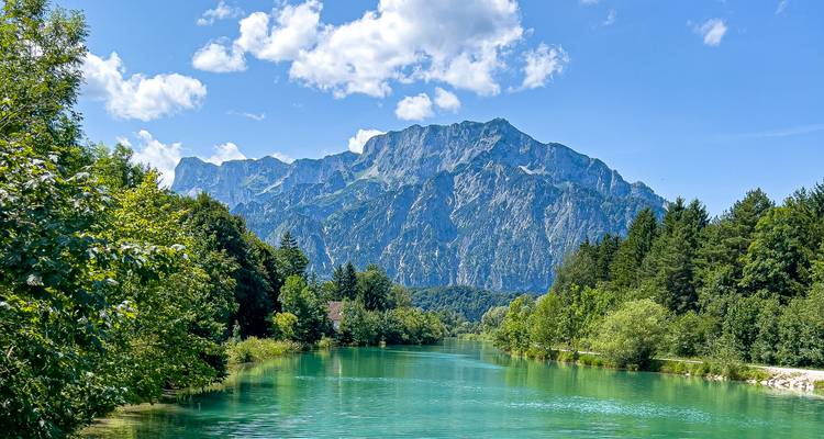 Crystal clear turquoise river flanked by lush forest with a towering limestone mountain under a bright, cloud-dotted sky.