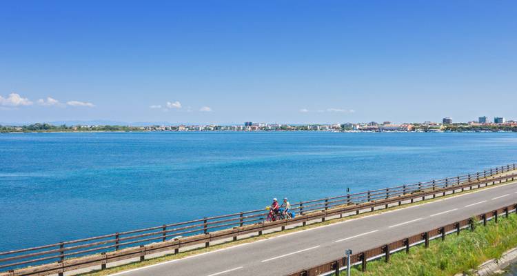 Cyclists pedal along a coastal causeway beside a vast blue lagoon under a cloudless sky.