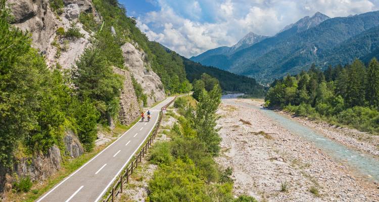 Cyclists follow a winding mountain road beside a stony riverbed surrounded by dense forest.