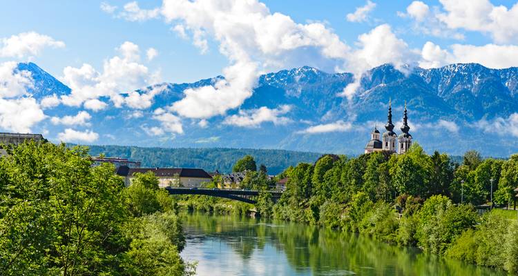 Vue panoramique d'une rivière avec un pont et une église. Des montagnes enneigées sont visibles à l'arrière-plan.