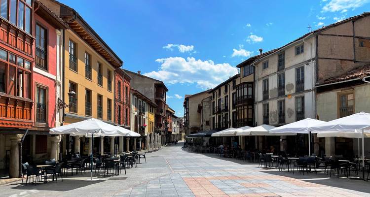 Une place de ville animée avec des bâtiments colorés et des terrasses de restaurants, sous un ciel bleu éclatant.
