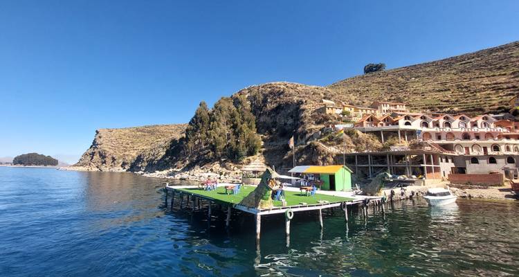 Een kustgezicht met een drijvend platform met groen, een steiger, en gebouwen op een heuvel naast een kalm water.