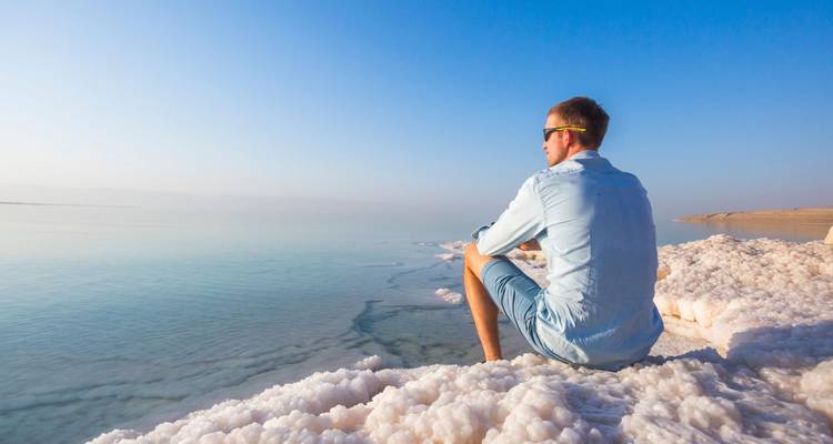 Mann in legerer Kleidung sitzt am salzkrustenbedeckten Ufer des Toten Meeres und blickt bei Tagesanbruch über das ruhige Wasser.