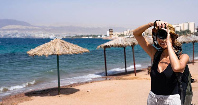 Weibliche Reisende macht Foto am Sandstrand mit Strohsonnenschirmen und Rotem Meer im Hintergrund.