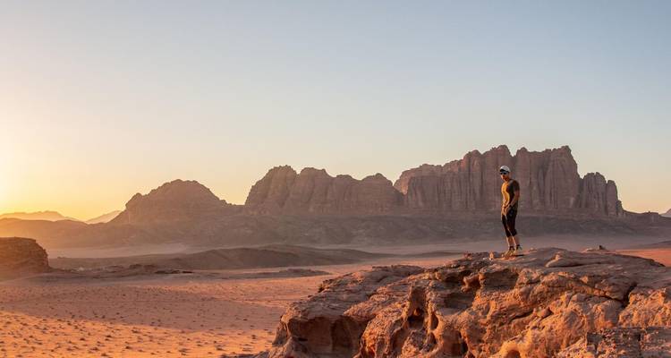Reisender steht bei Sonnenaufgang auf einem Felsvorsprung und blickt über die dramatischen Sandsteinberge des Wadi Rum.