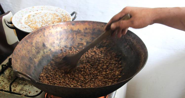 Une main remue des grains de café en torréfaction dans une grande poêle rustique au-dessus d'une flamme nue.