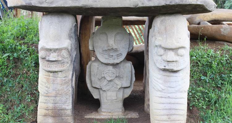 Des statues de pierre sculptées avec des visages et des figures anthropomorphes se dressent sous une dalle de pierre entourée d'herbe verte.