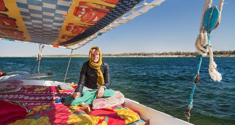 Une femme est assise en tailleur sur une felouque aux coussins colorés naviguant sur une rivière d'un bleu profond sous un ciel dégagé.
