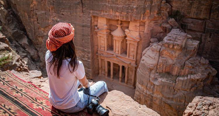 Depuis un tapis au sommet d'une falaise, une femme contemple la façade sculptée du Trésor taillée dans le canyon rose-rouge de Pétra.
