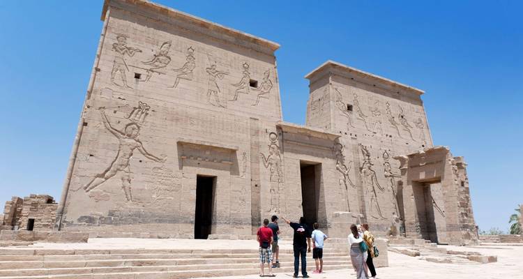 Des visiteurs se tiennent devant les imposants pylônes de grès d'un ancien temple égyptien couverts de reliefs sculptés sous un ciel bleu éclatant.