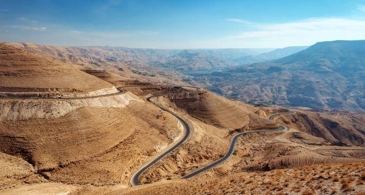 Une route d'asphalte sinueuse serpente à travers de vastes canyons arides et des collines accidentées sous un ciel bleu dégagé.