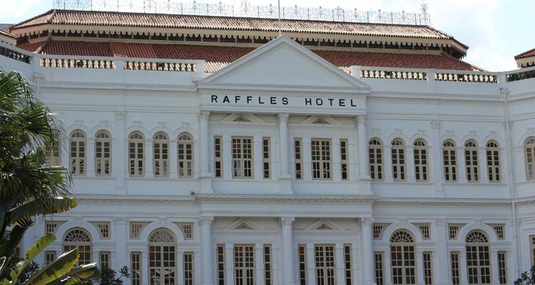 Élégante façade du Raffles Hotel d'époque coloniale avec des piliers blancs et des rangées de fenêtres à volets encadrées de verdure.