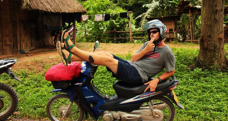 Voyageur détendu allongé de côté sur une moto bleue dans un cadre rural verdoyant, casque sur la tête et prenant la pose.