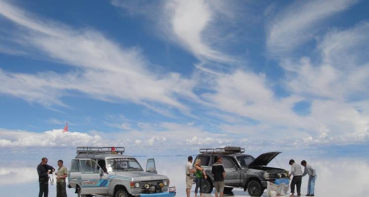 Off-road voertuigen en reizigers staan op de spiegelachtige zoutvlaktes onder een hemel doorstreept met witte wolken.