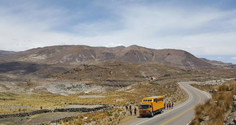 Gele overland expeditietruck gestopt op een kronkelende hooggebergtweg met reizigers die het kale berglandschap bewonderen.