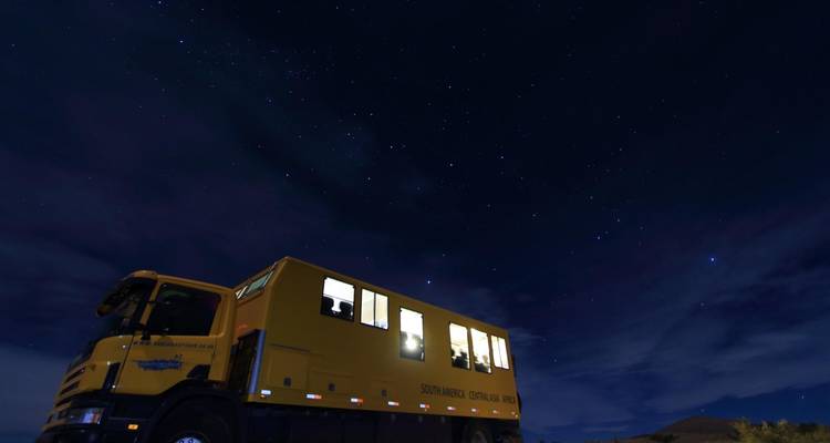 Sterrenrijke nachtelijke hemel torent uit boven een helder verlichte gele expeditietruck geparkeerd in de wildernis.