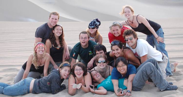 Grote groep lachende reizigers verdringt zich samen op golvende zandduinen voor een speelse foto.