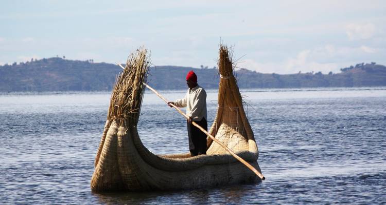 Een man peddelt een traditionele gevlochten rietboot over glinsterende meerwateren met heuvels op de achtergrond.