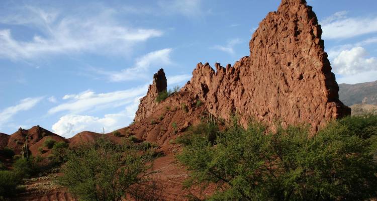 Grillige roodachtige rotsformaties rijzen op uit een schrale landschap onder verspreide wolken en blauwe lucht.