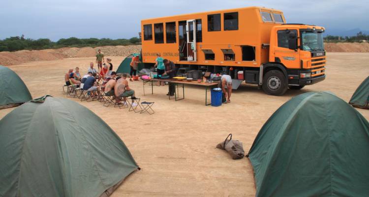Oranje expeditievrachtwagen geparkeerd naast opgezette groene tenten waar reizigers samenkomen voor een maaltijd in de buitenlucht op een stoffig veld.